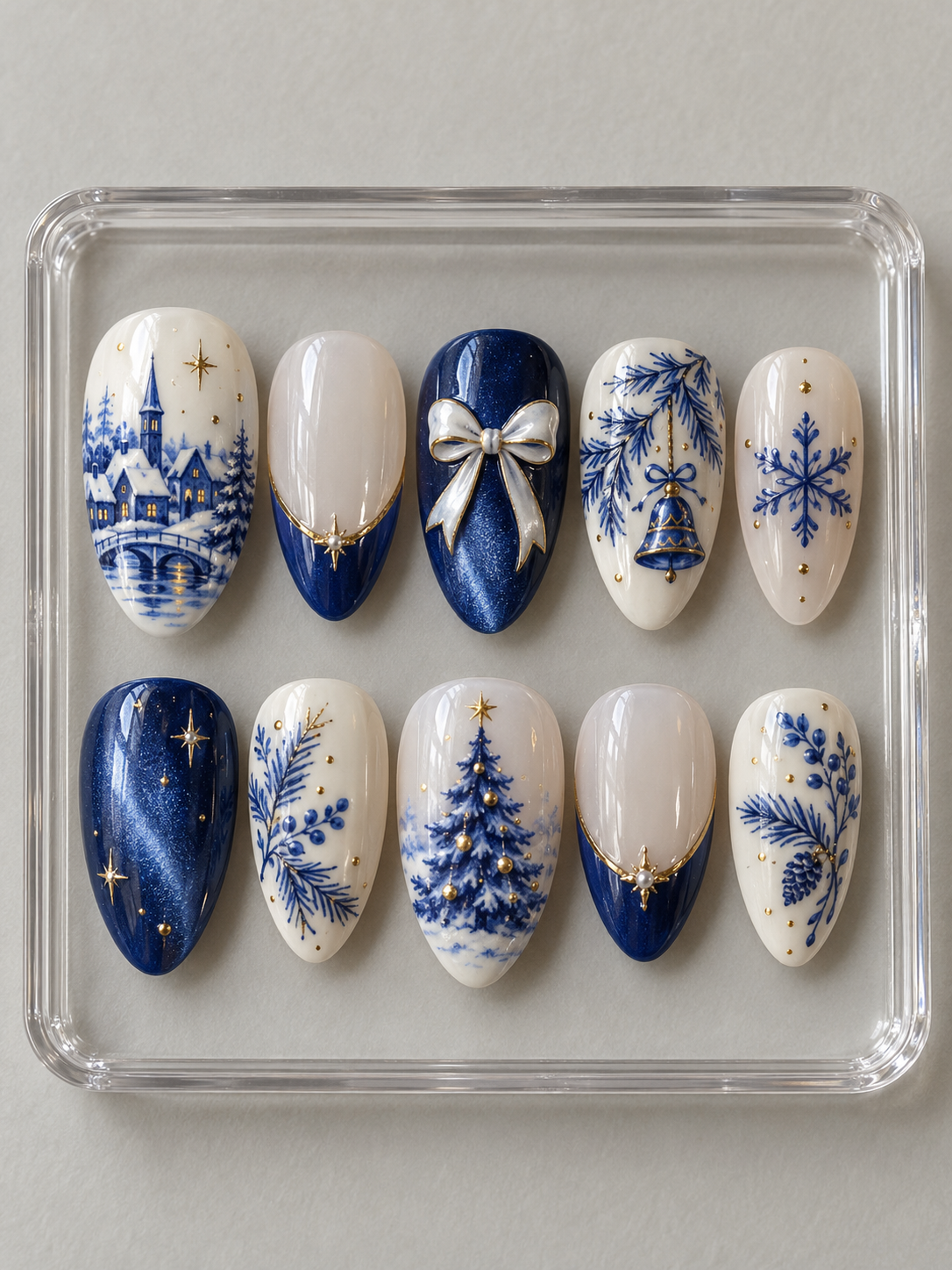 Ten cobalt blue and white Christmas press-on nails with snowy village, tree, botanical sprigs, bell, snowflake, white bow and gold-colour trim.